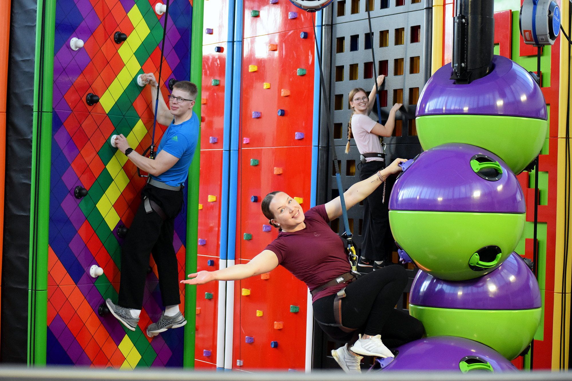 Three adults in harnesses climbing a rainbow wall and a purple-and-green ball obstacle